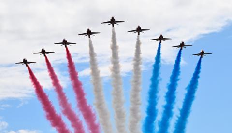 The Red Arrows flying in formation in the shape of an arrow head photographed from below. Three on the left have red smoke pouring from their tails, the middle three have white smoke and the three on the right have blue smoke. The sky is a pale cloudy blue.