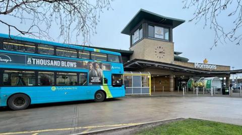 A blue double decker bus at a bus stop outside Morrisons supermarket entrance