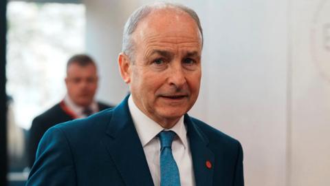 Micheal Martin wearing a blue suit, white shirt and blue tie. He has grey hair and is looking at the camera.