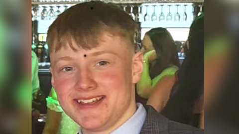 Kerr Paterson has short, brown hair and is smiling at the camera. He is wearing a blue shirt and a tweed suit jacket. He is in a bar with people standing behind him