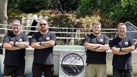 Four men wearing matching black T-shirts with a round white logo on, stand in front of a rowing boat with their arms folded. In the middle of them is a sign that has a rowing boat with four oars in a circle that reads "DeadLegz" 