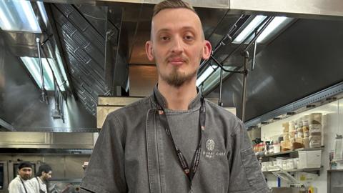 A man stands in a kitchen. He has short brown hair and facial hair. He is wearing a grey denim shirt with a logo on it. In the background are several chefs with stripey black and white aprons on