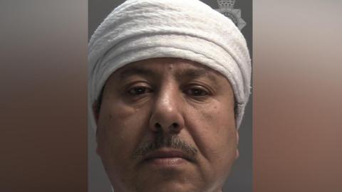 A custody picture of a man with a moustache who is wearing a white head covering. 