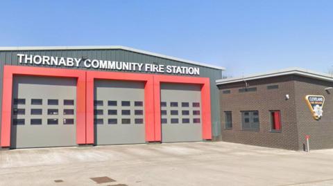Exterior of the fire station. It is a large single-storey garage with three large doors surrounded by red cladding; the words "Thornaby Community Fire Station" are written in large white doors above the doors. To the left stands a brown-brick building sporting the fire brigade's logo.
