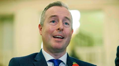 Paul Givan has short dark and grey hair and is speaking as the picture is taken. He has blue eyes and stubbled facial hair. He is wearing a black suit jacket. white shirt, dark blue tie and a red poppy on his lapel. 