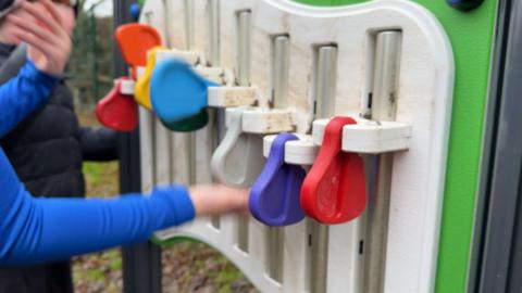 A child's hands are seen playing with a colourful toy. The equipment has plastic hammers, each in a colour of the rainbow, which can be flipped up and let go so they bang on white metal pipes.