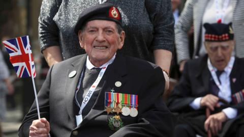 A veteran waves a Union Jack flag as he takes part in a VE Day Parade. He is wearing a suit with a line of medals across his chest.