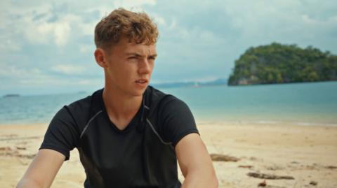 Alfie Watts sits on a beach with blue sea behind him. He is looking to the right of the camera and squinting in the sunshine. He is wearing a black T-shirt and has blond hair. 