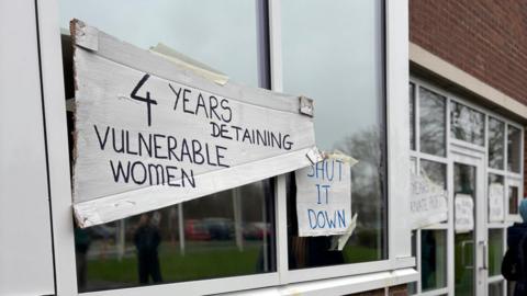 Close-up of windows of a detention centre. The windows are plastered with handwritten signs stating '4 years detaining vulnerable women' and "Shut it down'.