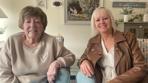 An image of Betty Unsworth and her daughter Gaynor Harrison. Betty is wearing a beige jumper with a white scribbled heart design, Gaynor has on a white blouse with a brown leather jacket. Both are sitting in blue chairs and are smiling. There is a painting of a woodland scene on the wall behind them.