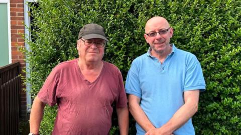 Two men standing next to each other in front of a bush. One is wearing a T-shirt and a hat and the other is wearing a polo shirt. They are both looking into the camera.