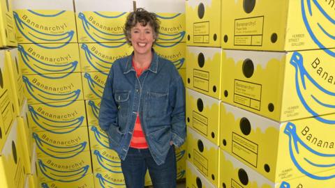 Lady smiling with short wavy hair, blue denim jacket, striped red and orange jumper and blue jeans. She is stood next to a stack of yellow boxes with Bananarchy written on them.