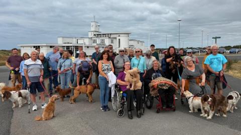 About 30 people, many with dogs, gather on Birkdale Common to protest plans to allow the R&A to take over a section of the public land ahead of the 2026 Open golf championships there. Three people in wheelchairs sit in the foreground, in front of the standing protesters. 