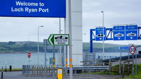 Signs for the Loch Ryan ferry port and services to Belfast sit above a roadway with a view out over the sea