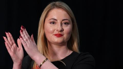 The Labour MP for Wellingborough and Rushden is seen standing just off centre in the image. She is clapping, and looking just past the camera. She has a black top on, blonde hair, and red nails and lipstick. 