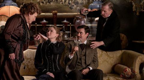 Some of the lead actors from Night and Day in a behind-the-scenes shot wearing costumes from the film. The character Jennifer Saunders is playing stands on the left and is leaning over two actors sitting on a sofa. The character played by Timothy Spall is standing on the right and holding up a glass 