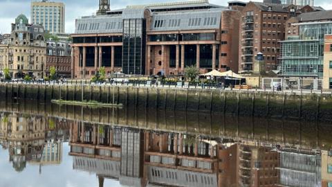 A large red court building reflected in a river running in front of it