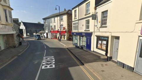 East Street in Okehampton on a sunny day. People are walking along the pavement in the distnace and a blue van is driving along the road. An estate agent called Bradley's which has blue hoardings is on the right hand side.