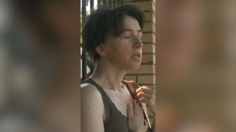 A woman with short hair stands in the street with her eyes closed and hand on her chest