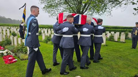 Six men in RAF suits carry a coffin draped in a Union Jack while another RAF man walks behind. They are in a cemetery