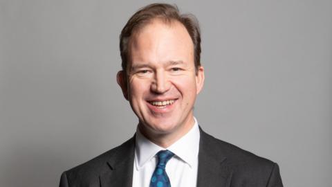 A portrait photograph of MP Jesse Norman. He is wearing a grey suit jacket and white shirt with a blue spotty tie. He is stood against a grey backdrop.