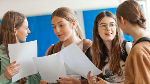 Four female students standing together in a classroom or school corridor, holding sheets of paper and talking in a small group. Blue lockers or notice boards are visible in the background, and the group appears to be reviewing or discussing the documents. 