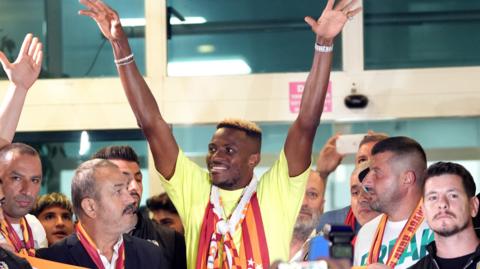 Victor Osimhen wears Galatasaray colours around his neck and raises both arms in the air as he is surrounded by fans on his arrival in Turkey