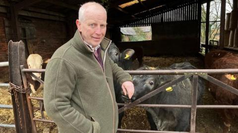 Richard Yates has very short white hair and is wearing a dark green zip jumper. He is leaning on a metal fence into a barn. Beyond the fence there are some curious black and brown cows near him, one is sniffing his hand. There is hay on the ground of the barn and a window in the back corner showing greenery and a farm building.