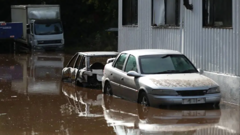 Three badly damaged vehicles sit in muddy flood water. 
Two cars are positioned near an outbuilding and a lorry sits at an angle.