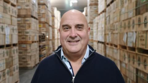 Richard Clothier in his cheese storage facility, wearing a navy zip jumper and a light coloured top beneath and smiling at the camera. There are boxes of cheese behind him. 