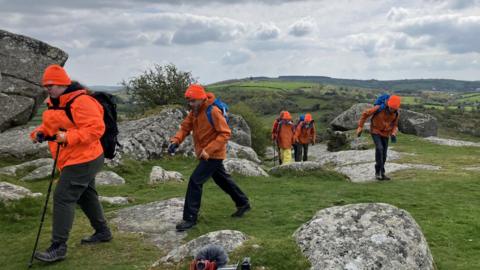 Young people wearing orange waterproofs and orange woolly hats are carrying rucksacks and walking across granite outcrops and grass.