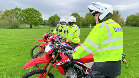 Three police officers wearing white helmets are each holding onto a red off-road bike. They are standing in a field with football posts and trees in the distance.