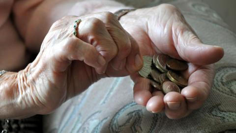 Close view of an elderly person's hands. The person is wearing a gold ring with green stones embedded in it, a silver bracelet on one hand and a silver watch on the other. They have a pile of coins in one hand and are holding a single coin in the other. One of the hands is resting on what looks like the armrest of a chair.