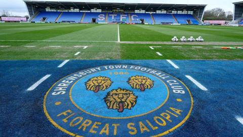 The Shrewsbury Town club crest with a stand at the Croud Meadow in the background