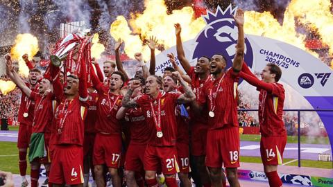 Liverpool Football Club celebrate winning the Premier League title. A group of players in red wearing medals jump in the air, lifting a silver trophy. In the background we can see the stadium, with fireworks and a banner that reads 'Champions 2024/25'.
