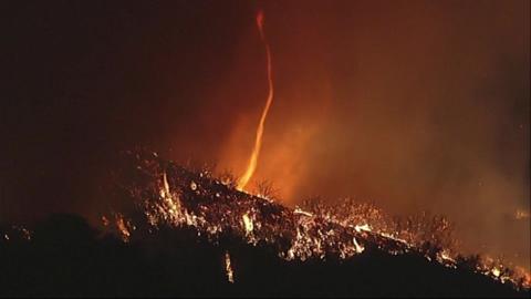 A funnel of fire burns upwards to dark smoke on a hillside at nighttime.