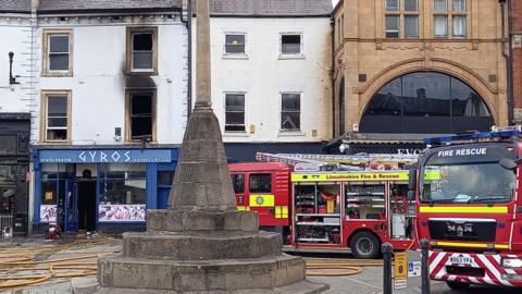 Two fire engines parked in a market place outside a row of shops and takeaways. 