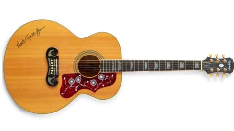 A brown acoustic guitar is pictured, with Noel Gallagher's signature seen on the body of the guitar. Around the sound hole is a detailed red pattern.