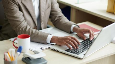 A close up of a business person in a brown suit and white shirt (we only see from the neck down) typing on a grey laptop on a desk. The desk has a white and red mug, a hole puncher and stationary.