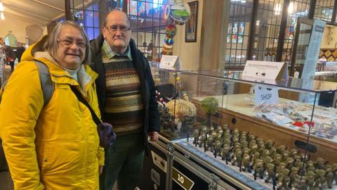 A woman in a yellow coat is stood next to a man in a striped jumper with a dark-coloured coat over the top. The pair are stood by a clear box which contains several rows of knitted, miniature World War Two soldiers.