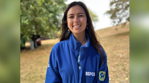 A young woman with long dark hair looks at the camera, she is standing in front of a tree. She has a nose piercing and earrings. She is wearing a blue RSPCA-branded fleece.


