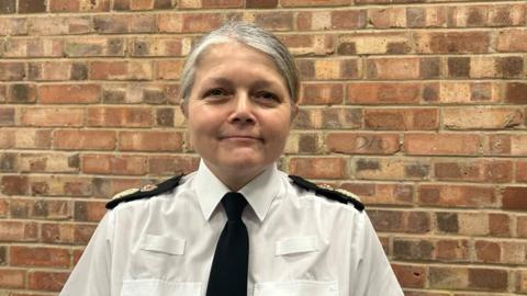 Standing in front of a bare brick wall, Sarah Crew smiles at the camera. She wears a white Avon and Somerset Police shirt and a black tie. 