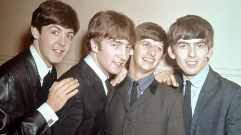 Paul, John, Ringo and George are wearing black, brown and grey suits and stand close together in the image. They have short cropped hair and are all smiling at the camera but are looking in different directions.