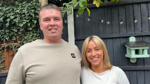 Kevin and Jodie stood side by side with their arms around each other and smiling. Kevin is wearing a beige jumper, and Jodie is wearing a white long sleeved top. The couple are stood in their garden, with a grey wooden fence behind them that's got fairy lights strung along the top of it. 
