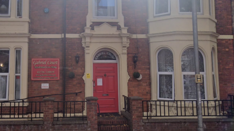 A red brick terraced building with a red door and a red sign for Gabriel Court Care Home.