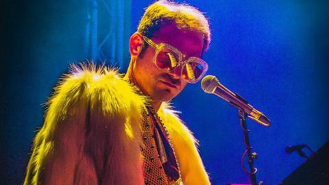 A performer in large sparkling glasses and a fluffy yellow coat stands beside a microphone under bright blue stage lights. The close-up image captures the atmosphere of a live music performance.