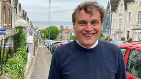 56-year-old Peter May is looking directly at the camera and smiling. You can see his teeth. 
He has wavy hair and a round face. 
Peter May is standing on a street that is on a hill with a view of the sea behind. 