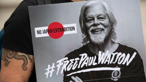 A demonstrator holds a portrait placard reading "no Japan extradition, free Paul Watson" during a demonstration in support of Paul Watson. Watson has white hair and a white beard and is smiling.