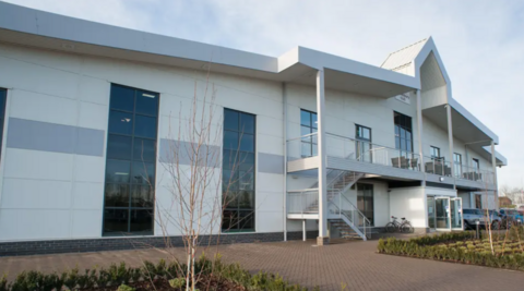 A large white building with tall tinted-glass windows and a large arch above the entrance and small shrubs on a paved area in the foreground