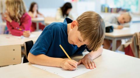 A young boy writing on a piece of paper, he has dark blonde hair and is wearing a navy polo shirt.